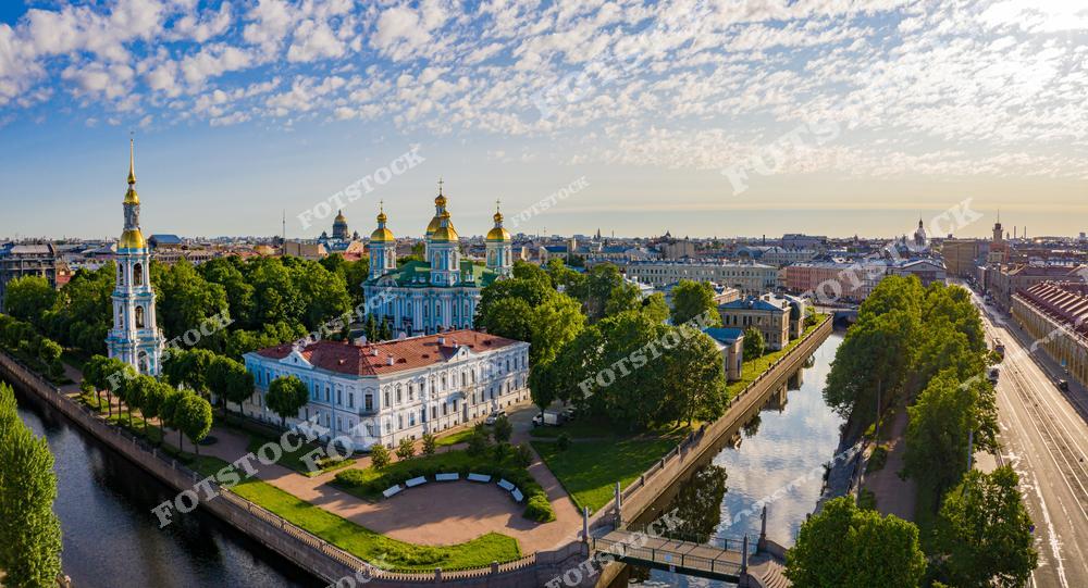На переднем плане изображены золотые купола Николо-Богоявленского морского собора и его колокольня, расположенные вдоль каналов Санкт-Петербурга. Рядом с собором проходит набережная с аккуратными зелеными насаждениями и дорожками. Летняя зелень в сочетании с исторической архитектурой создает спокойную атмосферу. На заднем плане можно увидеть панораму города с его известными достопримечательностями и зелеными улицами.